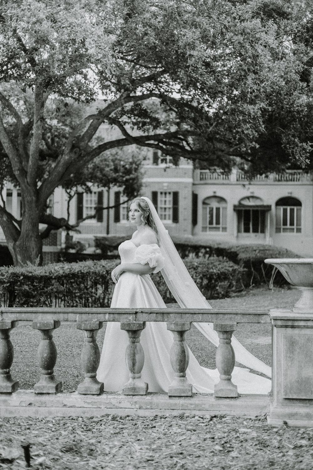 A classic black and white photograph shows a figure in a white wedding dress and veil standing by ornate stone balustrades.