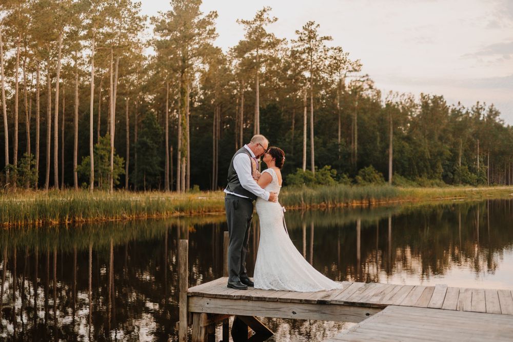 Couple embraces during sunset at a lakeside wooden dock with tall pine trees in the background.