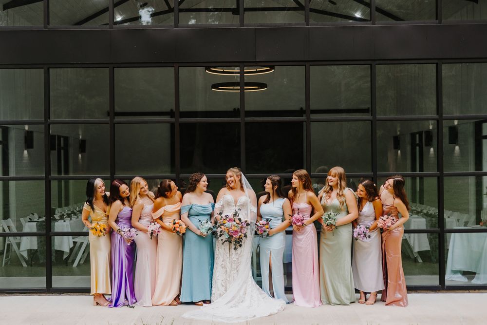 Wedding party members in pastel colored dresses stand together in a row outside a modern glass building.