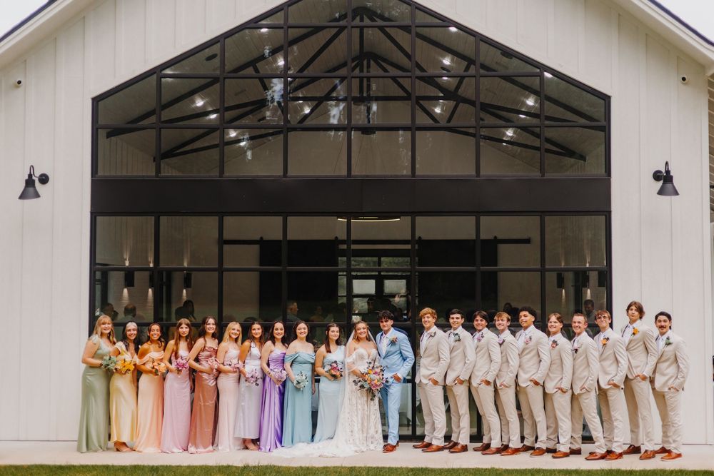 A wedding party in pastel dresses and suits stands in front of a white building with black trusses.
