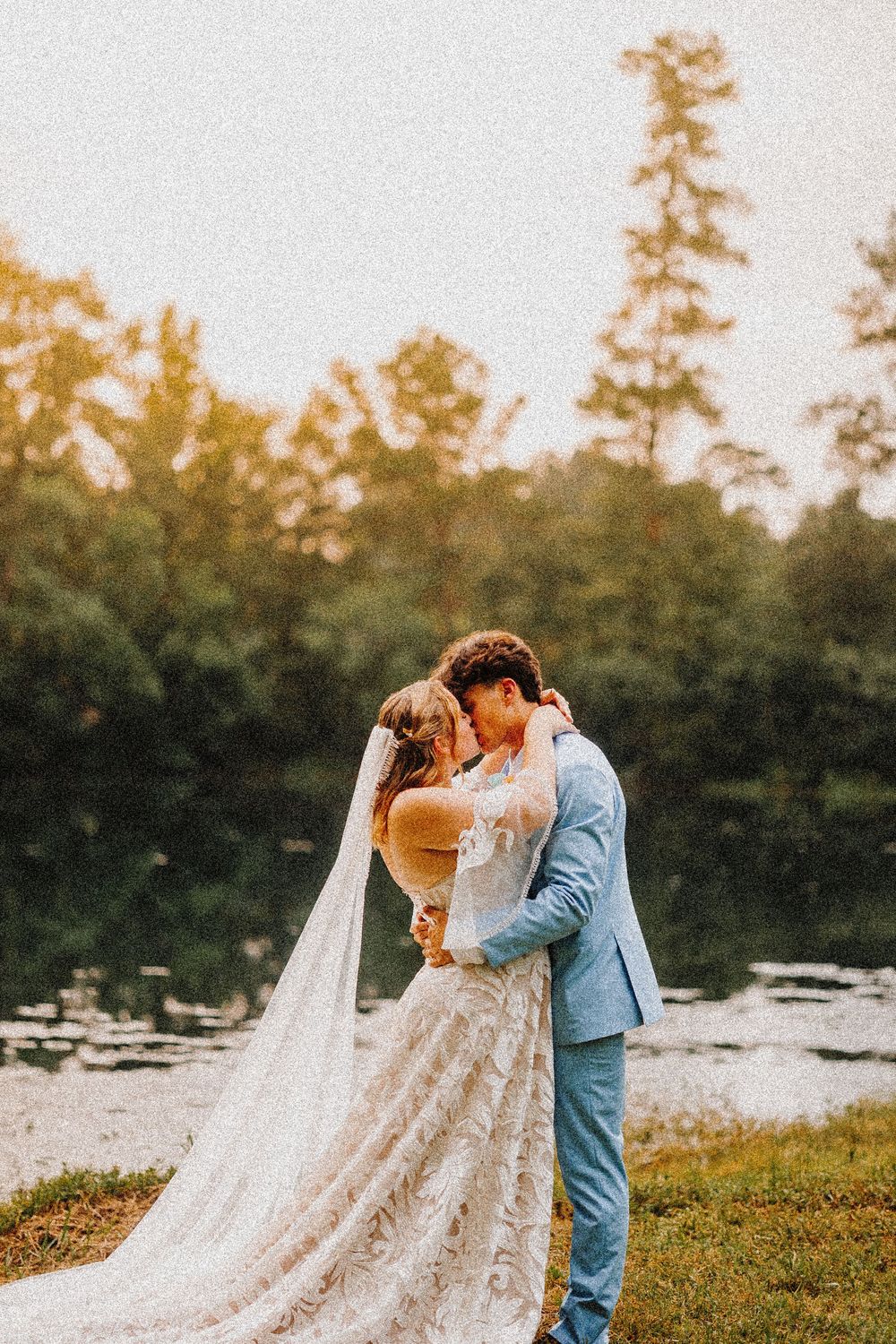 A romantic wedding couple embraces by a lake at sunset with a flowing lace veil and light blue suit.