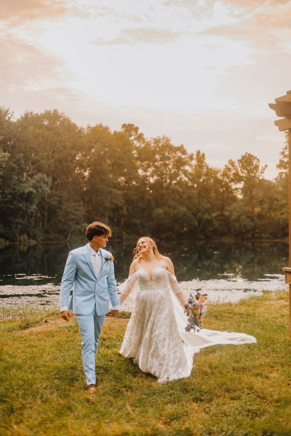 A couple in wedding attire walks through a sunlit meadow at golden hour with trees in the background.