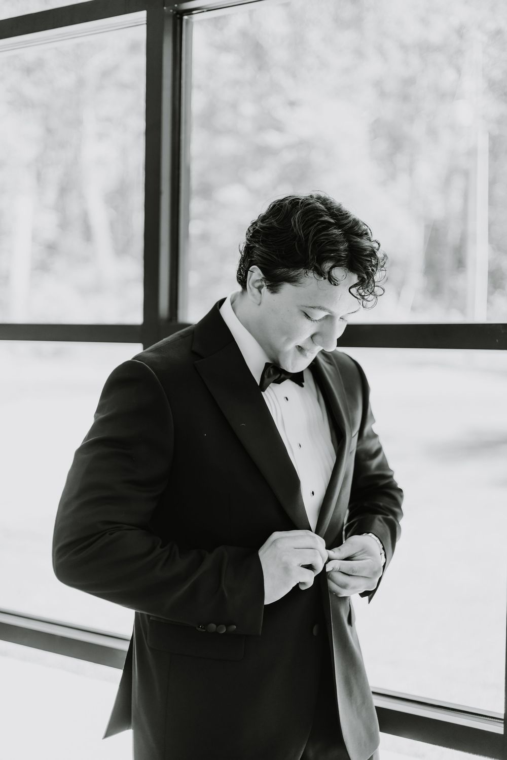 Black and white photo of a person in a tuxedo adjusting their bowtie while standing by large windows.