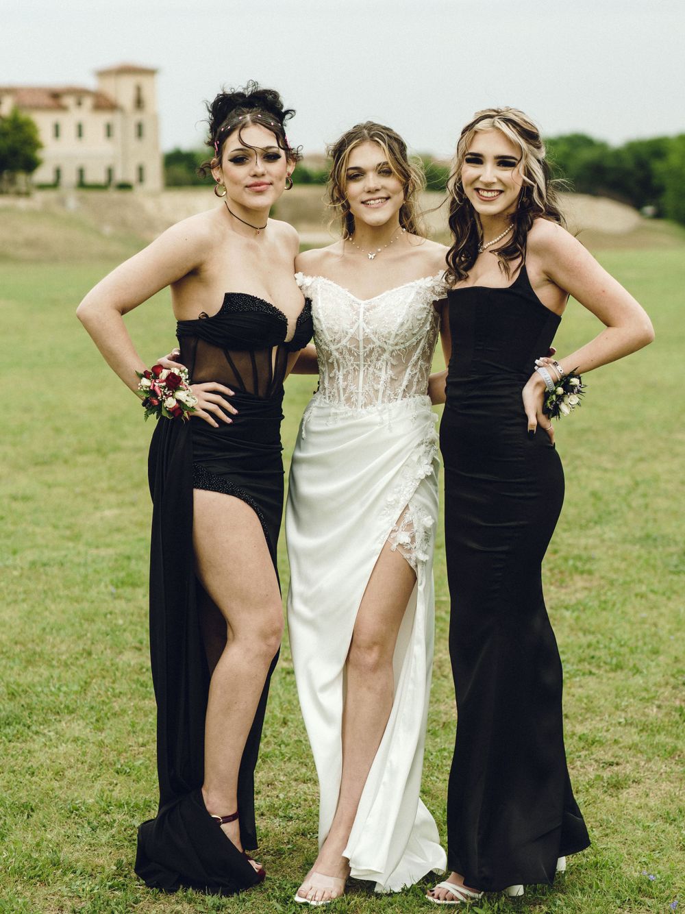Three women pose in formal dresses on a lawn, with the bride in white and bridesmaids in elegant black gowns.