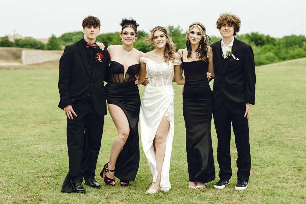 Five people in formal attire pose together outdoors on a grassy field wearing black and white dresses and suits.