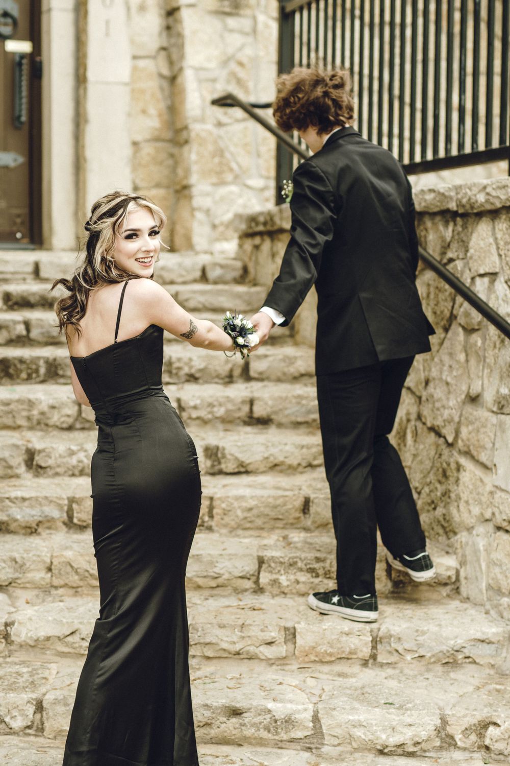 Elegant couple in formal attire ascending stone steps, with person in black gown looking back while holding drink.