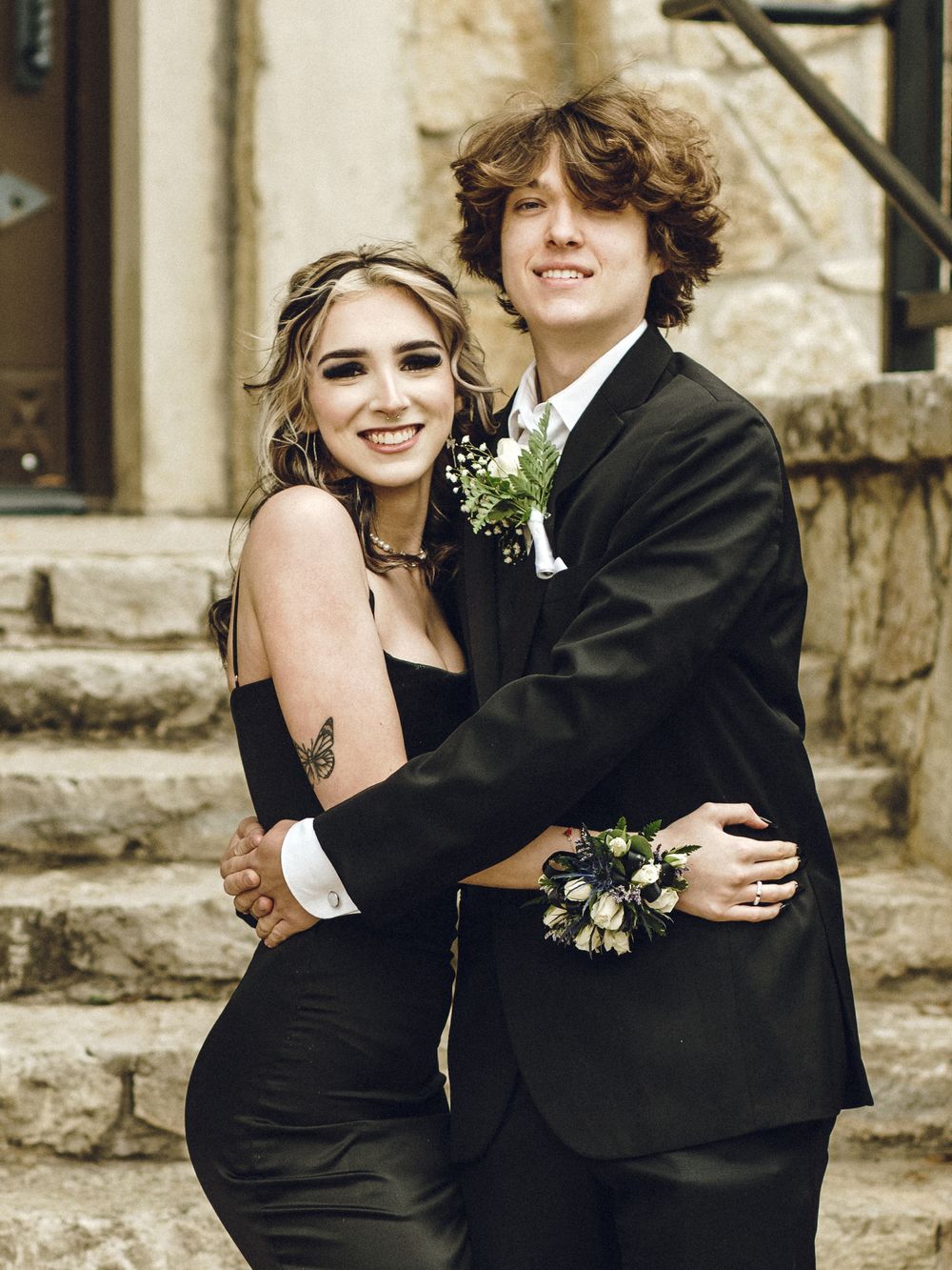 A couple dressed for prom or formal event embrace on stone steps, with the woman in black and man in traditional suit.