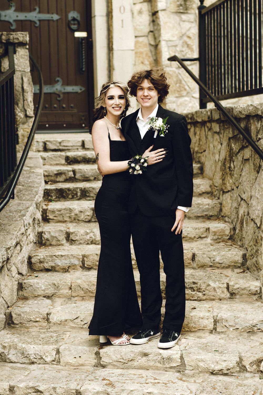 A couple in black formal attire poses together on stone steps outside a historic building for their prom or formal event.