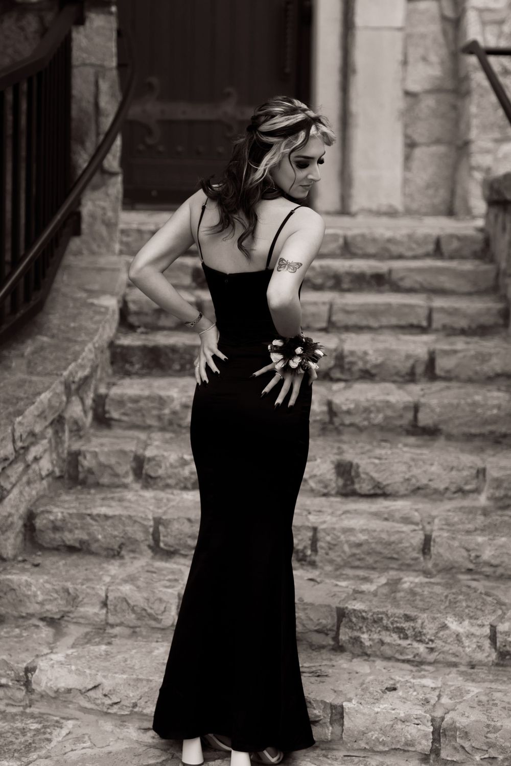 Woman in elegant black evening gown poses on stone steps in dramatic black and white vintage-style photograph.