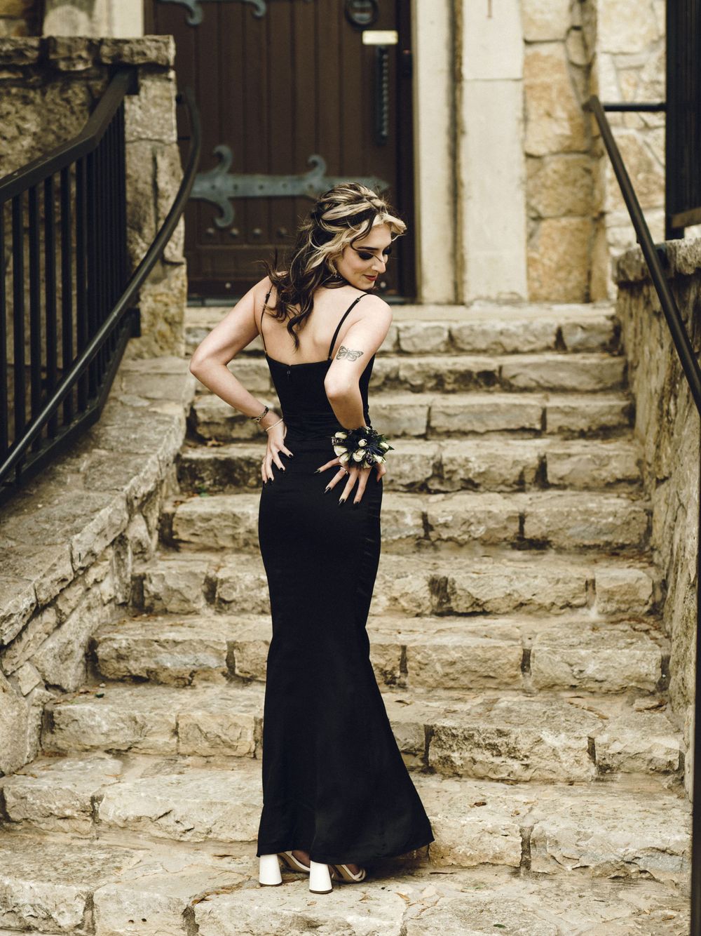 A person in an elegant black evening gown poses on stone stairs near rustic architecture.