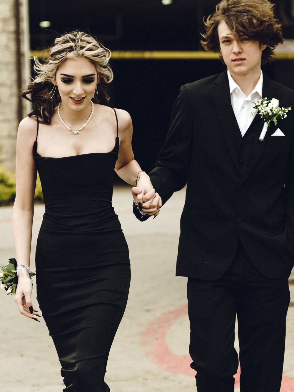 A couple in formal attire holding hands at prom, with her in a black dress and him in a classic black tuxedo.