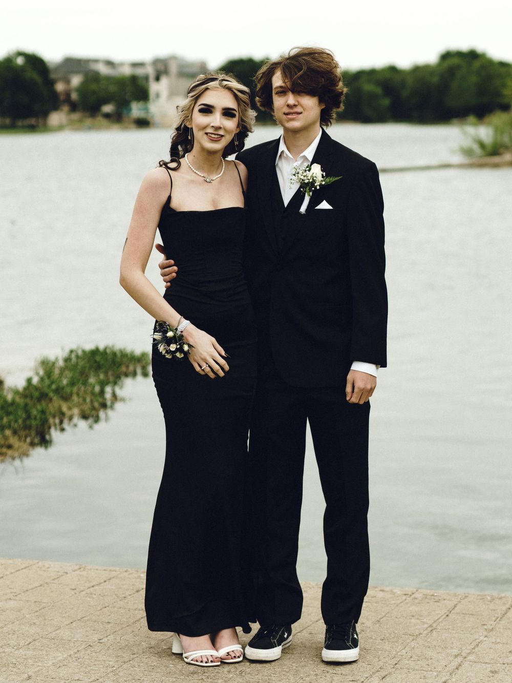 A formal couple poses together outdoors by a waterfront, wearing black formal attire for prom or a special occasion.
