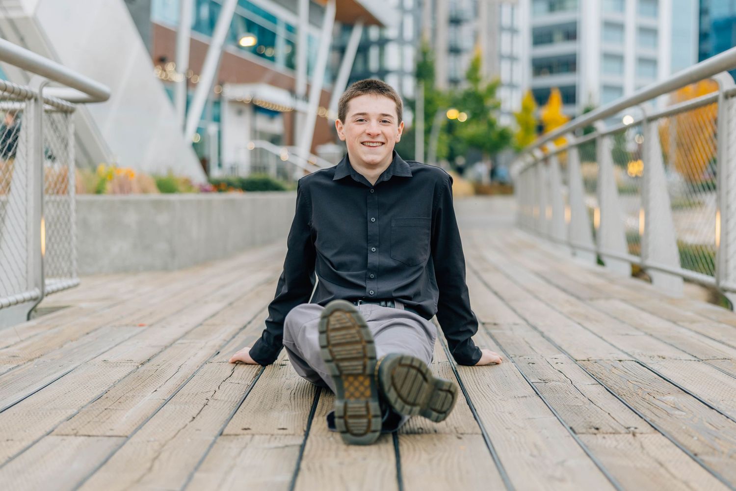 Smiling teenage boy in black shirt sitting cross-legged on a wooden bridge in an urban setting.