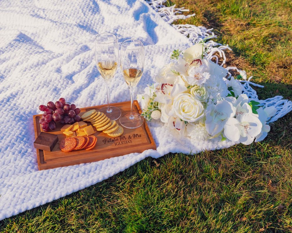 A picnic scene with wine glasses and appetizers arranged on a wooden board atop a white blanket on grass.