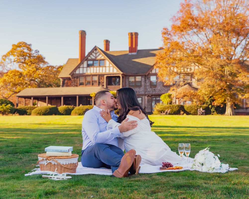 Romantic picnic on a lush green lawn with snacks and drinks in front of a grand estate during fall sunset.