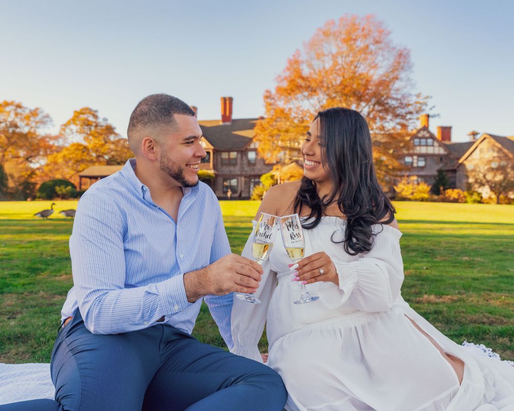 A couple shares champagne together while sitting on the lawn of an estate during autumn with fall foliage in background.