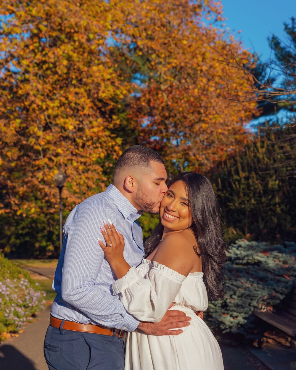 A happy couple embraces and smiles together on an autumn day with golden foliage in the background.