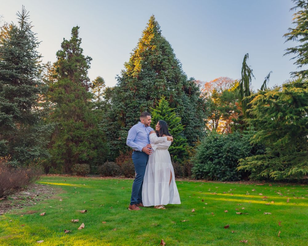 A couple shares a romantic moment on a grassy lawn surrounded by evergreen trees and golden sunlight.