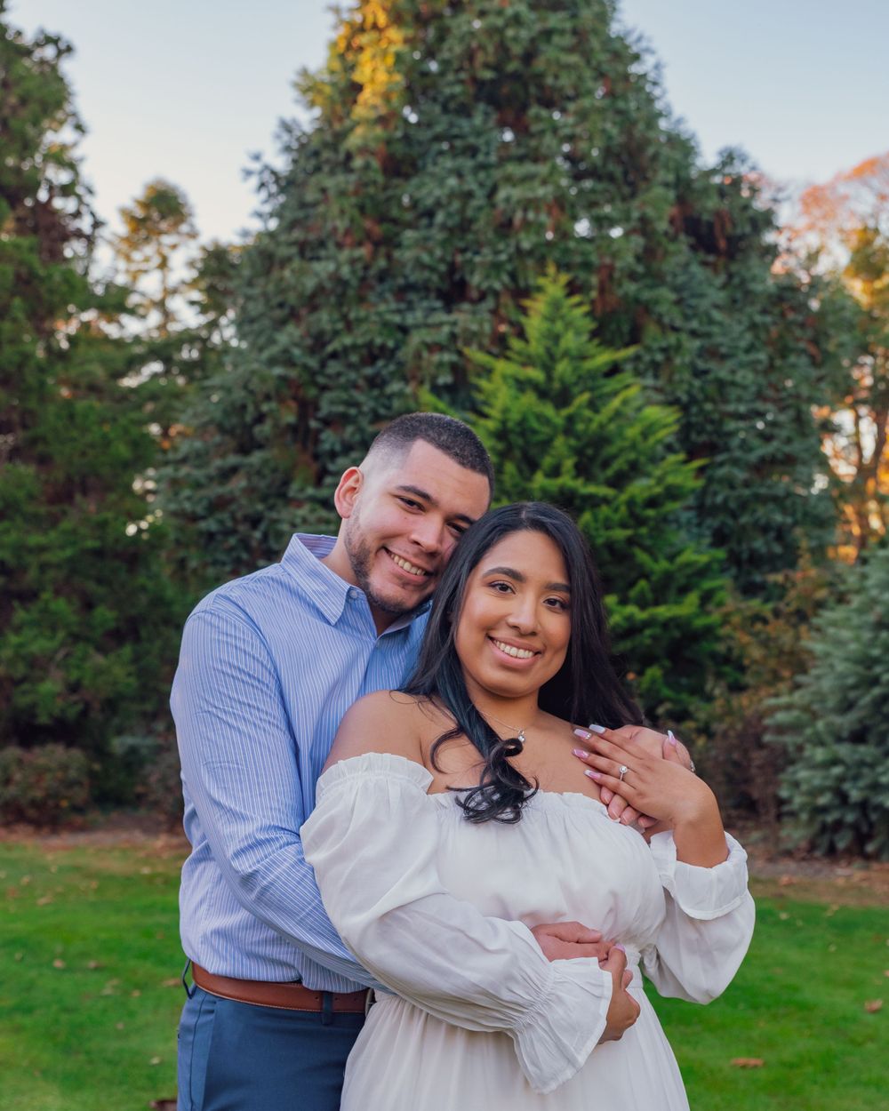 A couple embraces in a romantic outdoor portrait with evergreen trees and autumn foliage in the background.