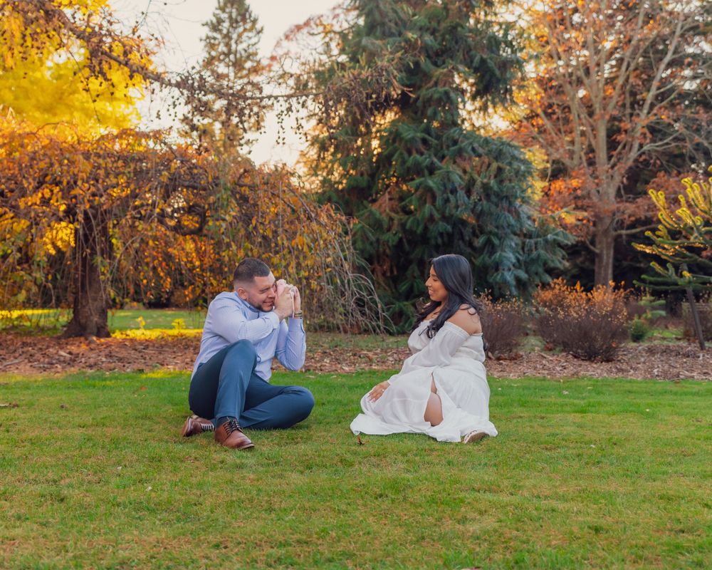 A couple sits together on autumn grass with colorful fall foliage and evergreen trees in the background.