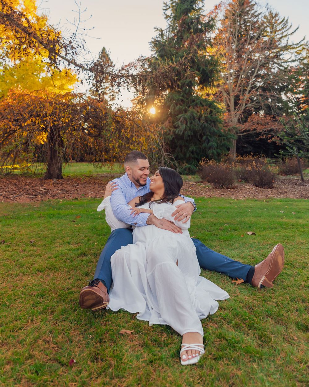 A couple sits on grass during golden hour, sharing an intimate moment surrounded by fall trees.
