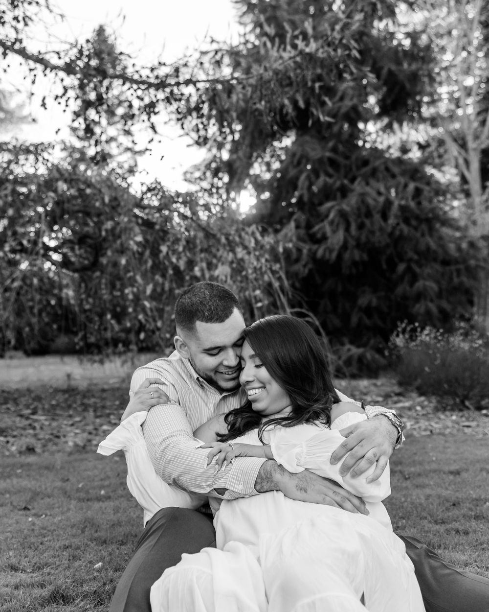 Black and white photo of a couple cuddling on grass beneath towering evergreen trees.