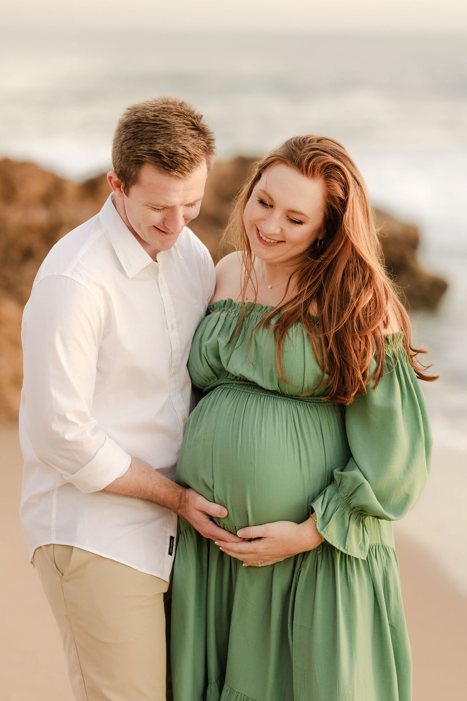 Red head mom and dad holding pregnant belly in a maternity pose at the beach