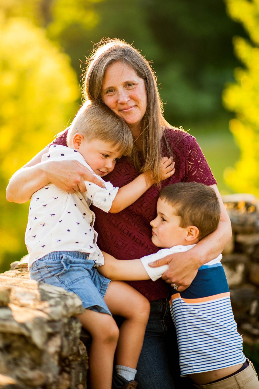 A mother smiles warmly while her two young sons embrace her outdoors in golden sunlight.