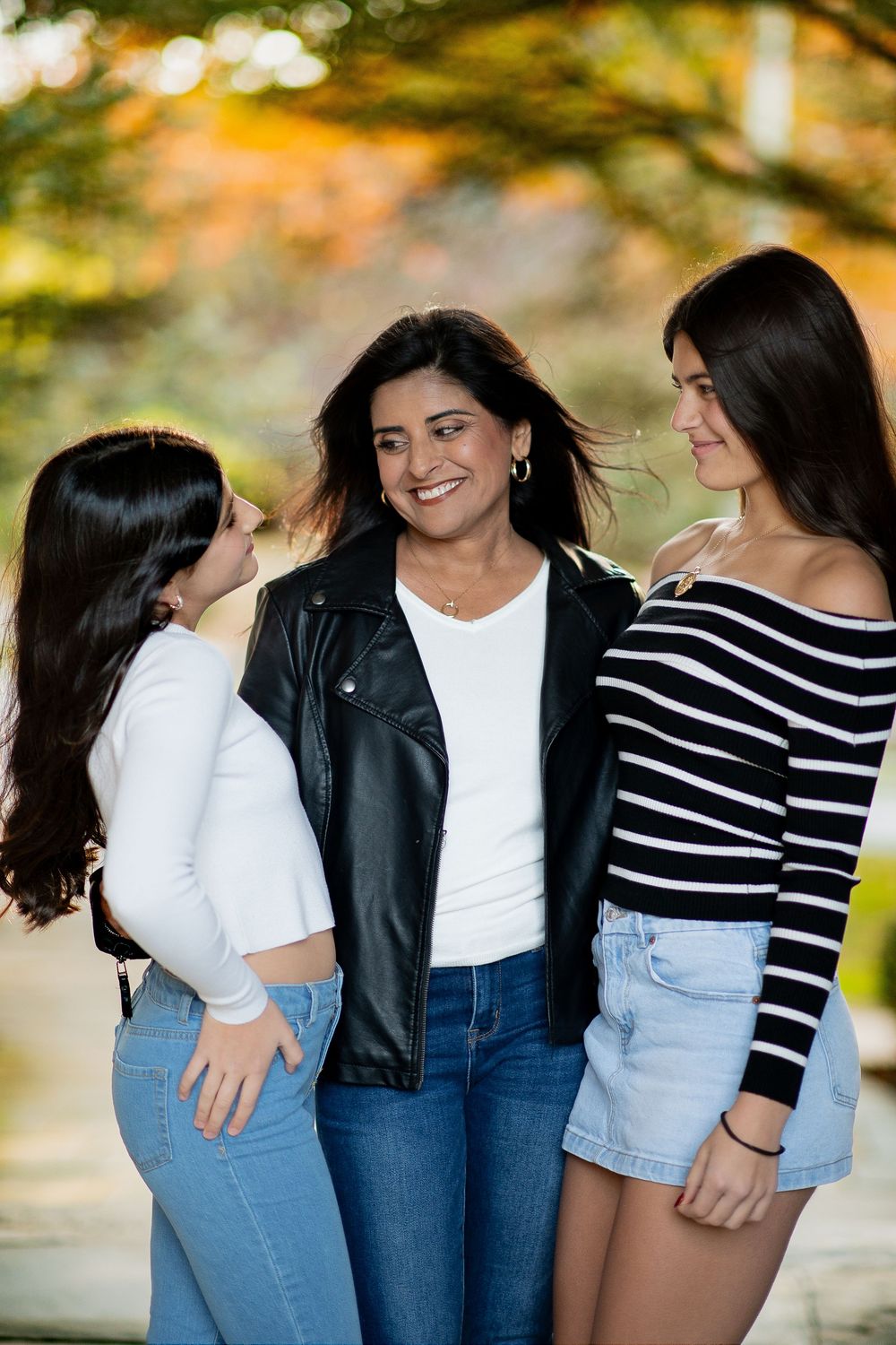 Three women smiling together outdoors in autumn, wearing casual outfits including jeans and a leather jacket.