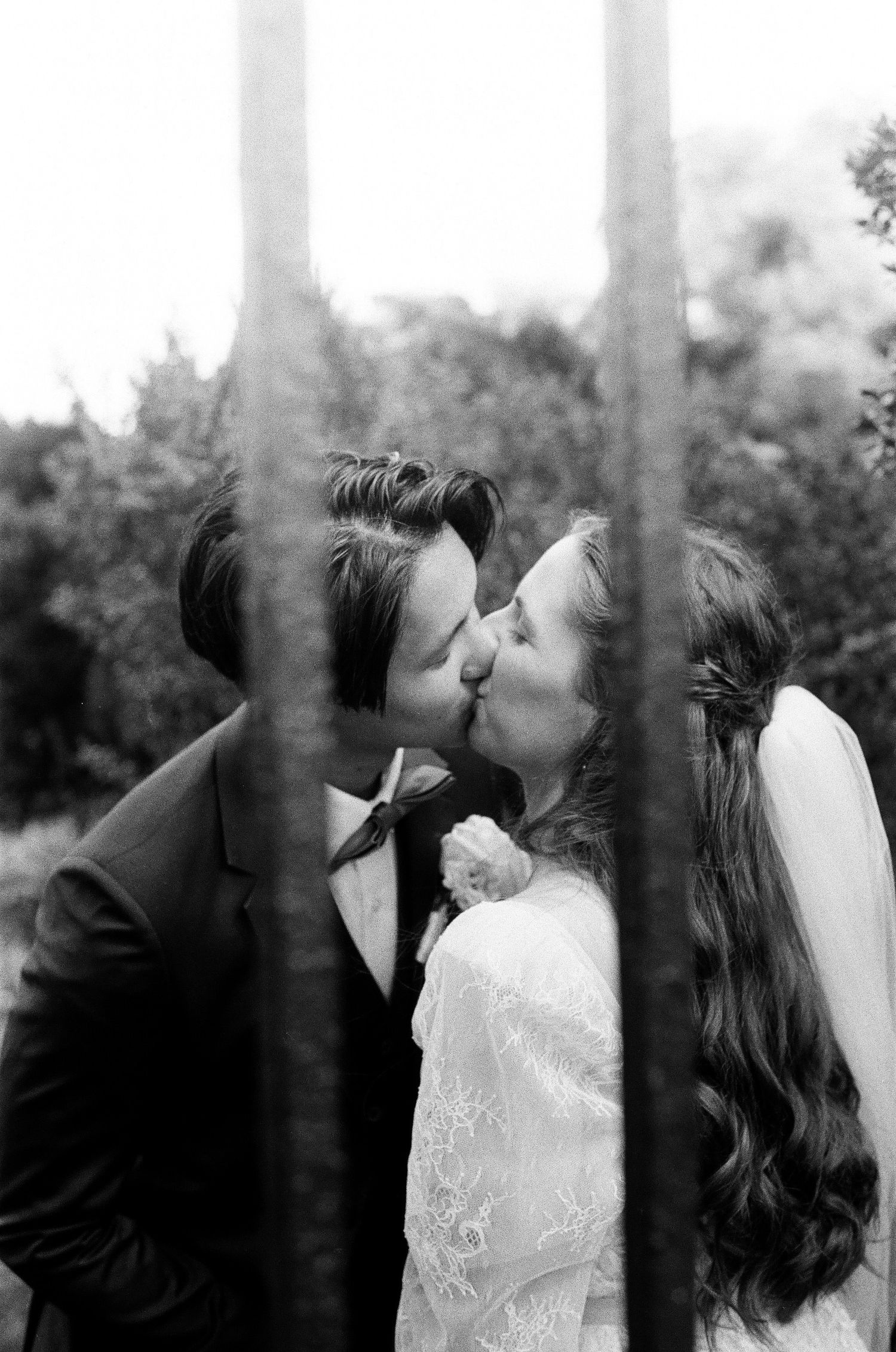 Black and white film photo of bride and groom sharing a kiss through trees on wedding day.