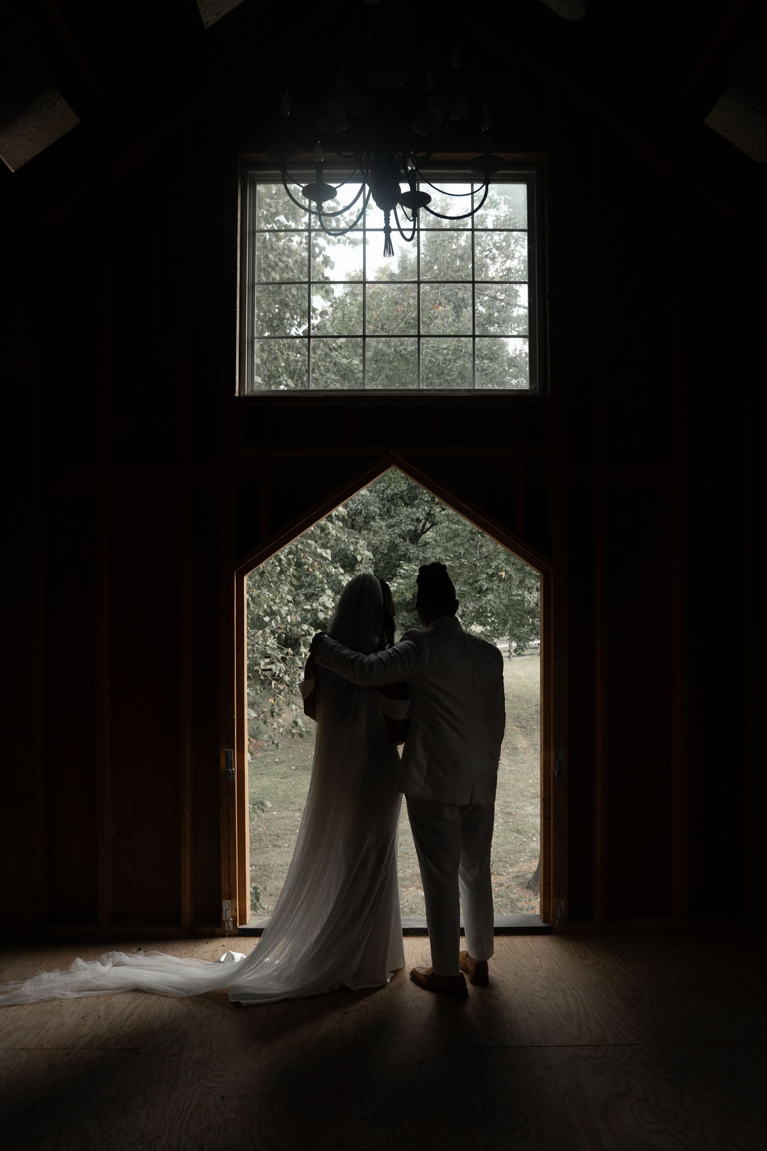 Silhouette of bride and groom embracing before arched window in dark rustic barn venue.