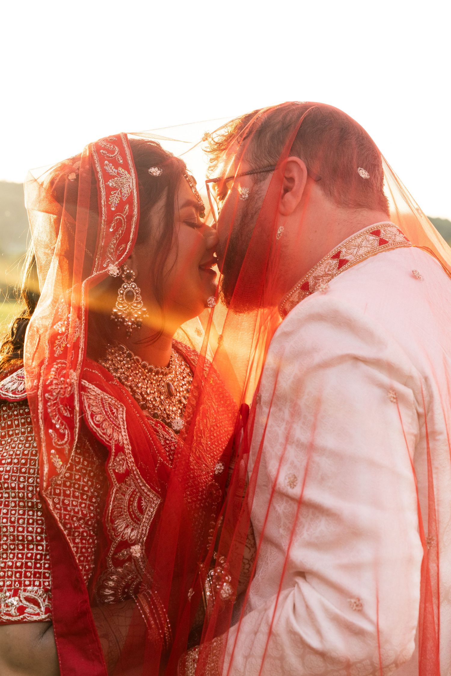 Indian bride and groom sharing a kiss under red bridal veil in golden sunset light.