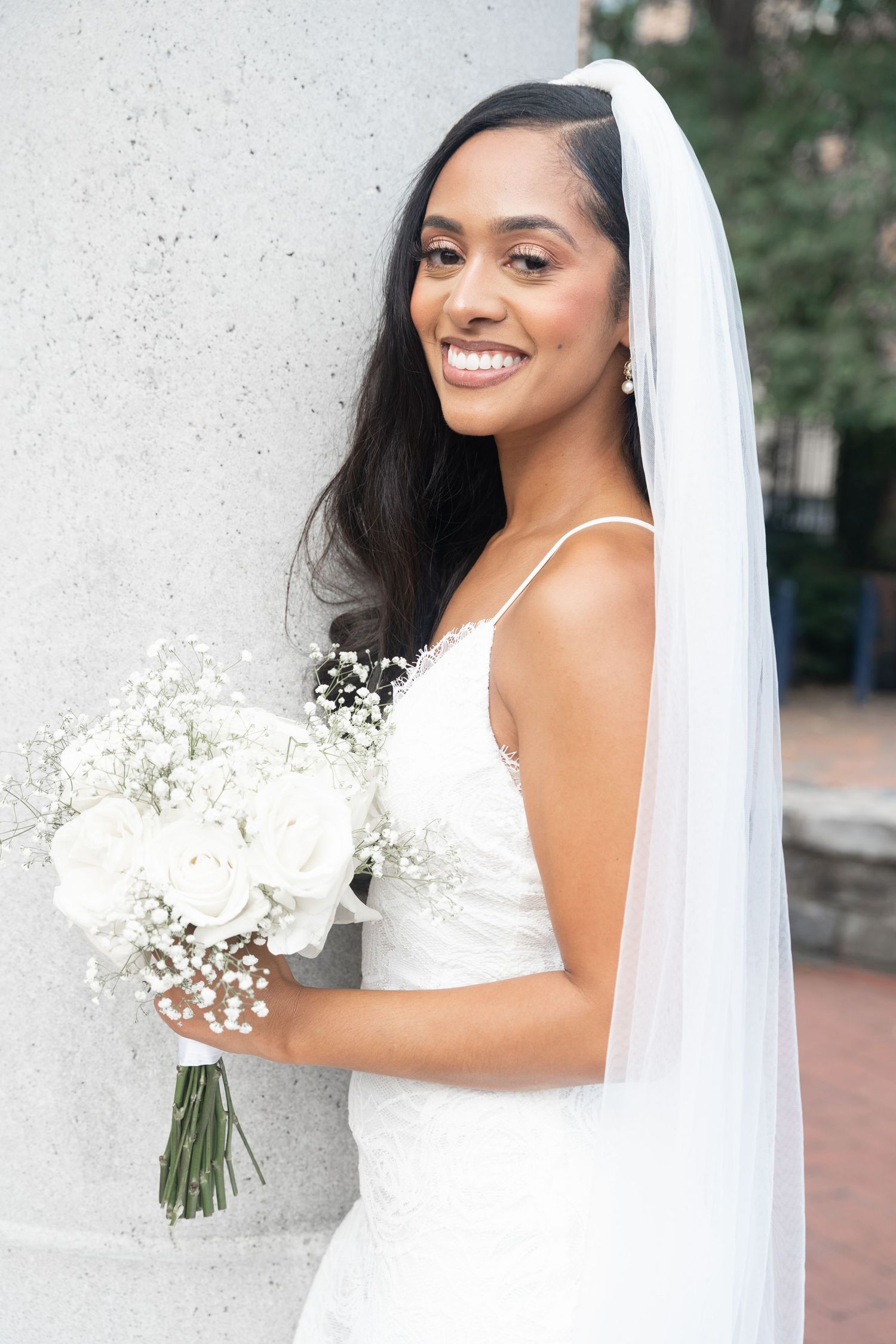 Radiant bride in white lace gown and veil holds a bouquet of white roses and baby's breath.