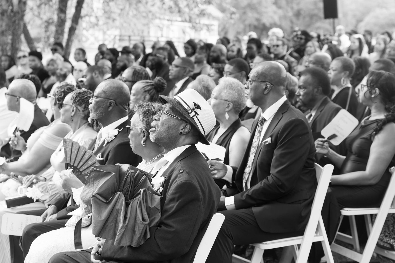 Black and white photo of diverse wedding guests seated and smiling during outdoor ceremony.