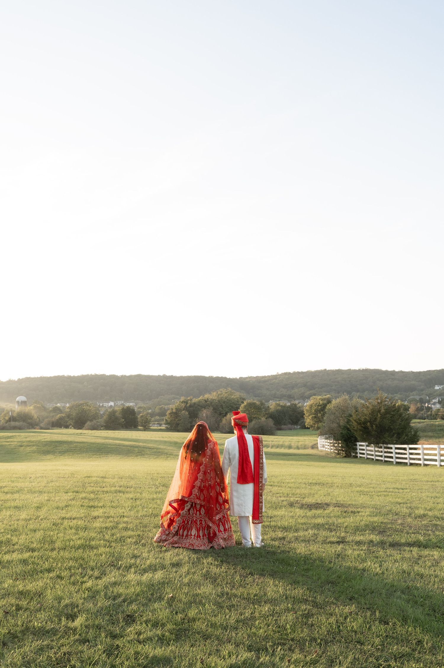 Indian bride and groom in traditional red and white attire walking through a scenic green field at sunset.