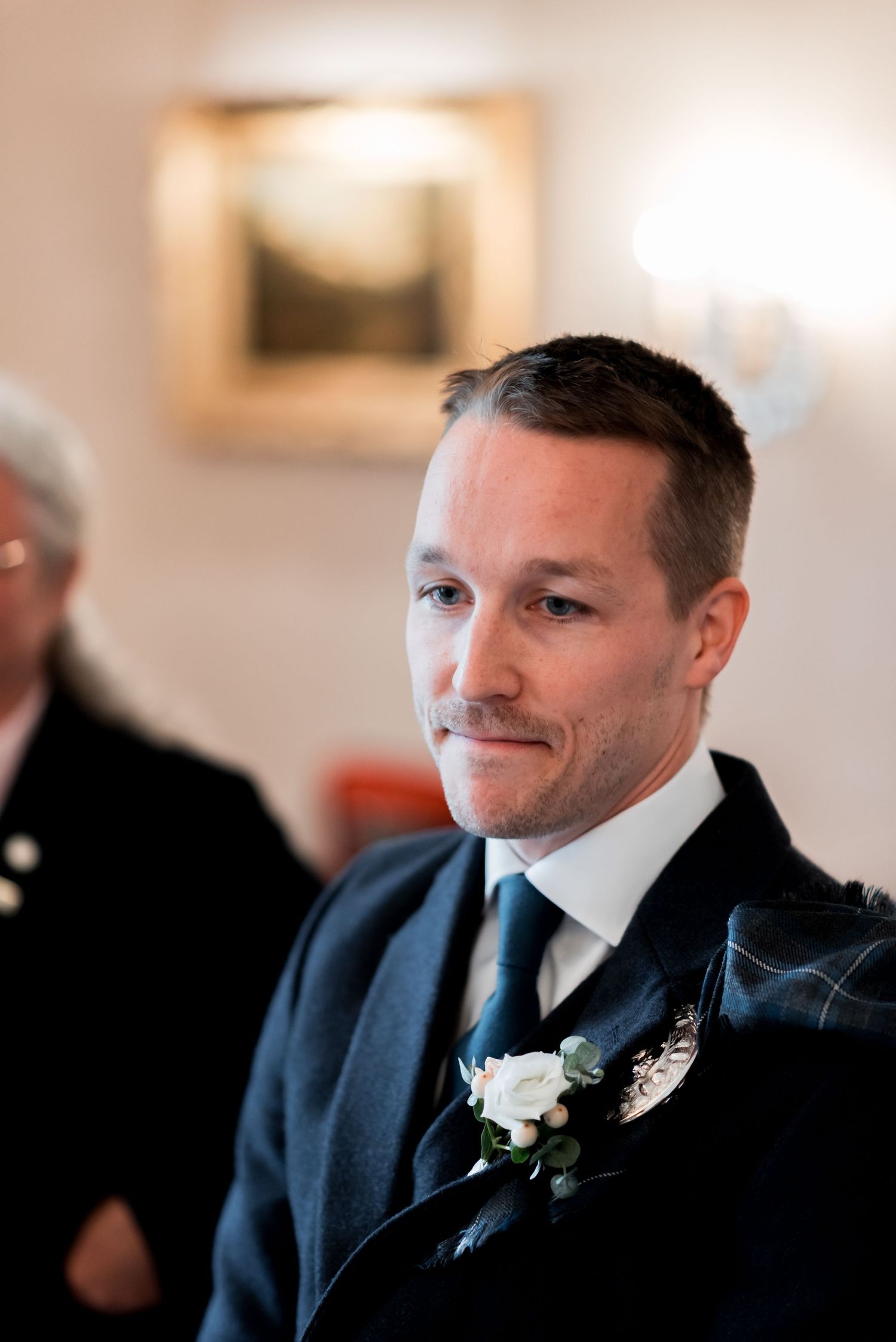 A groom in a navy suit with a blue tie and white boutonniere smiles during a wedding ceremony.