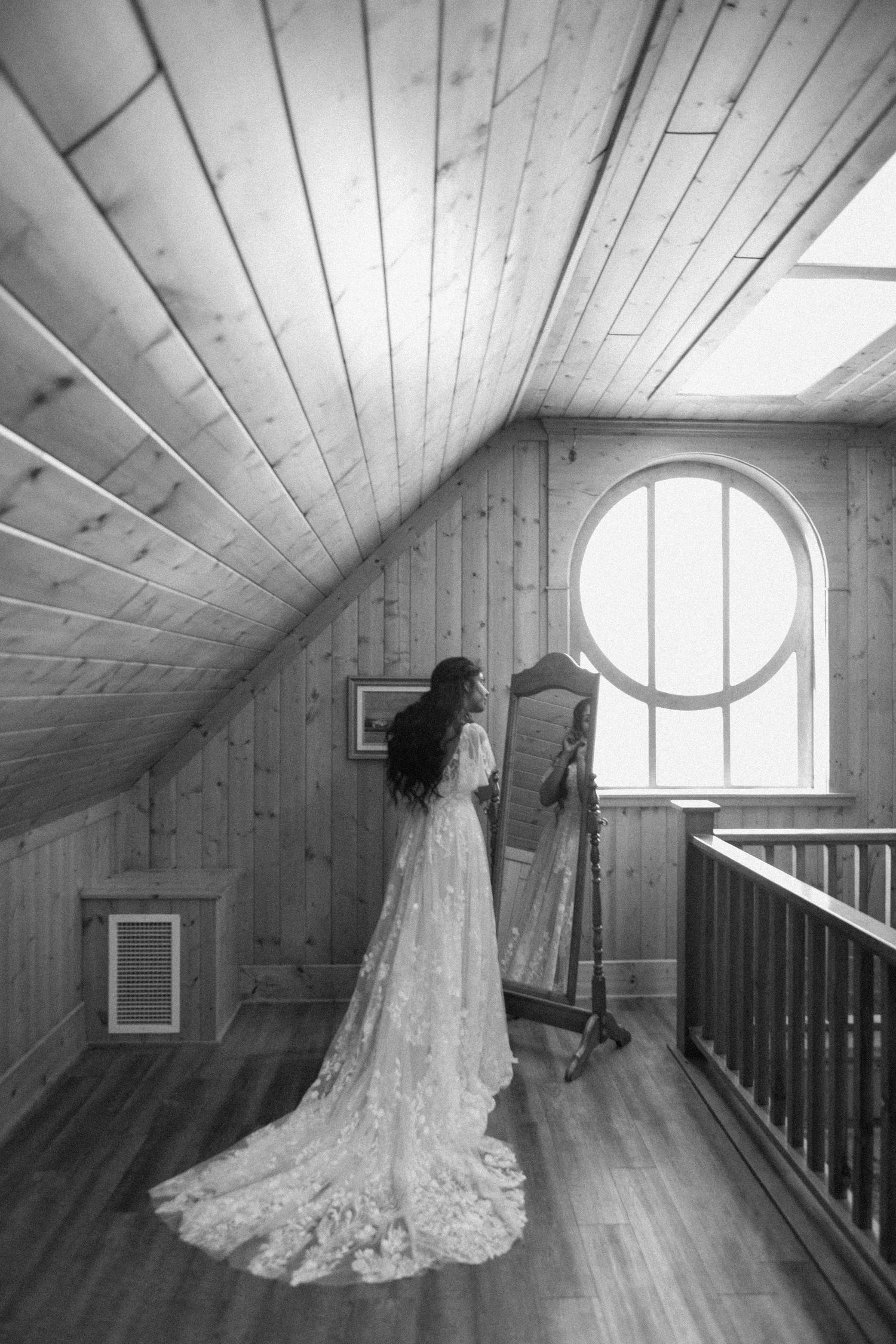 A person in a flowing lace wedding dress stands before a round window in a rustic attic room with wooden walls.