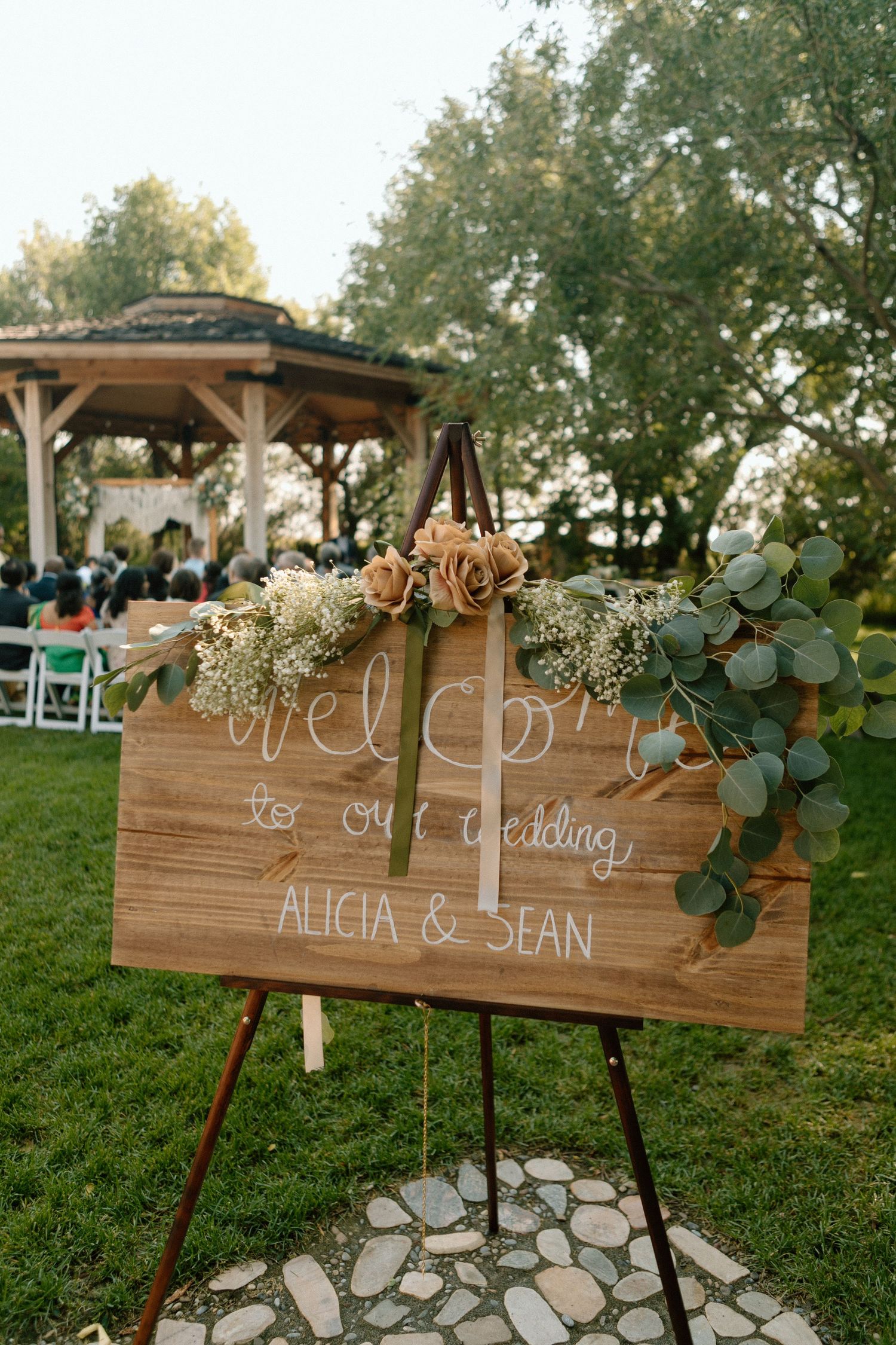 Rustic wooden wedding welcome sign decorated with eucalyptus leaves stands on an easel in an outdoor garden setting.