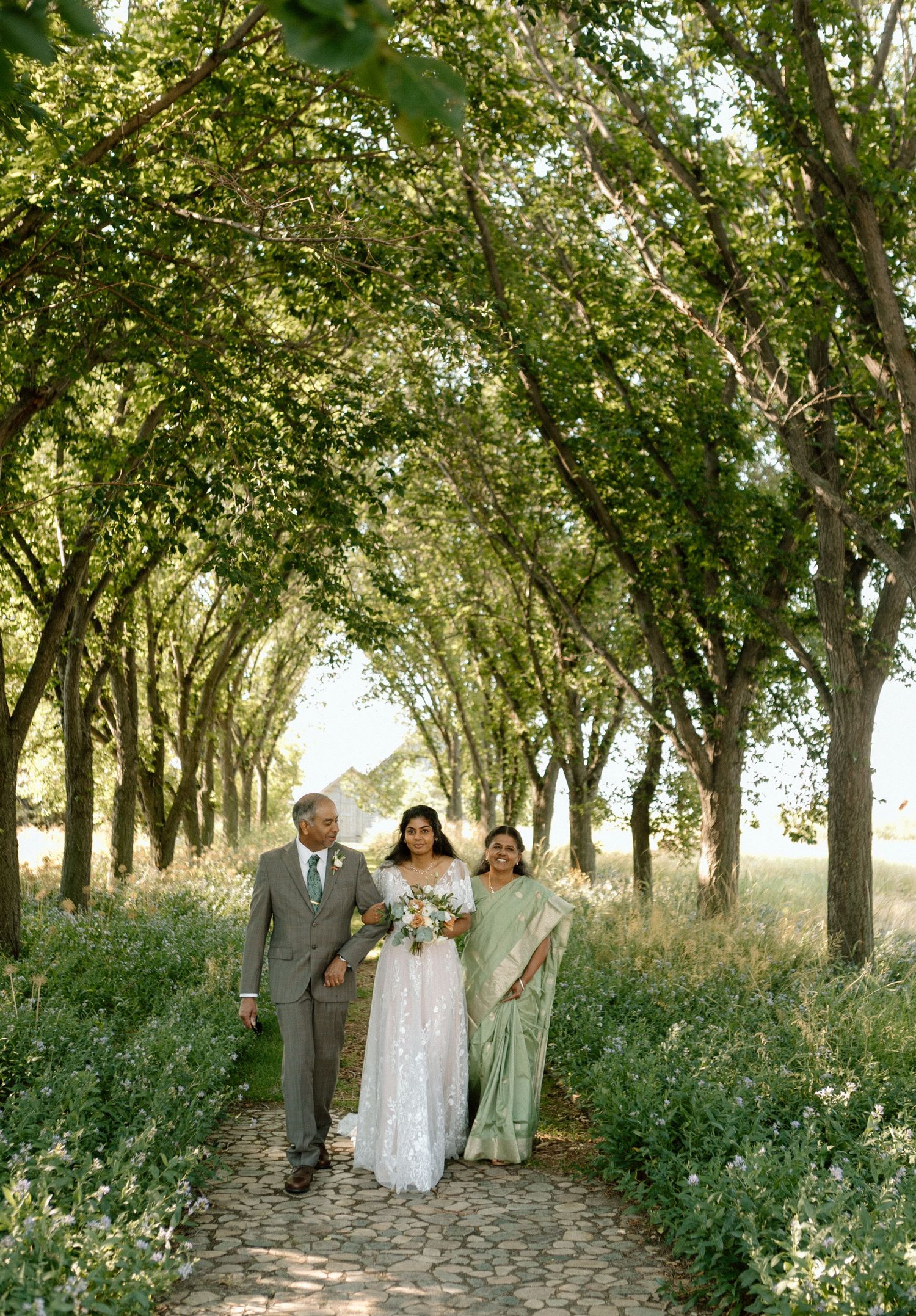 A tree-lined path with sunlight filtering through branches as wedding guests walk together outdoors.