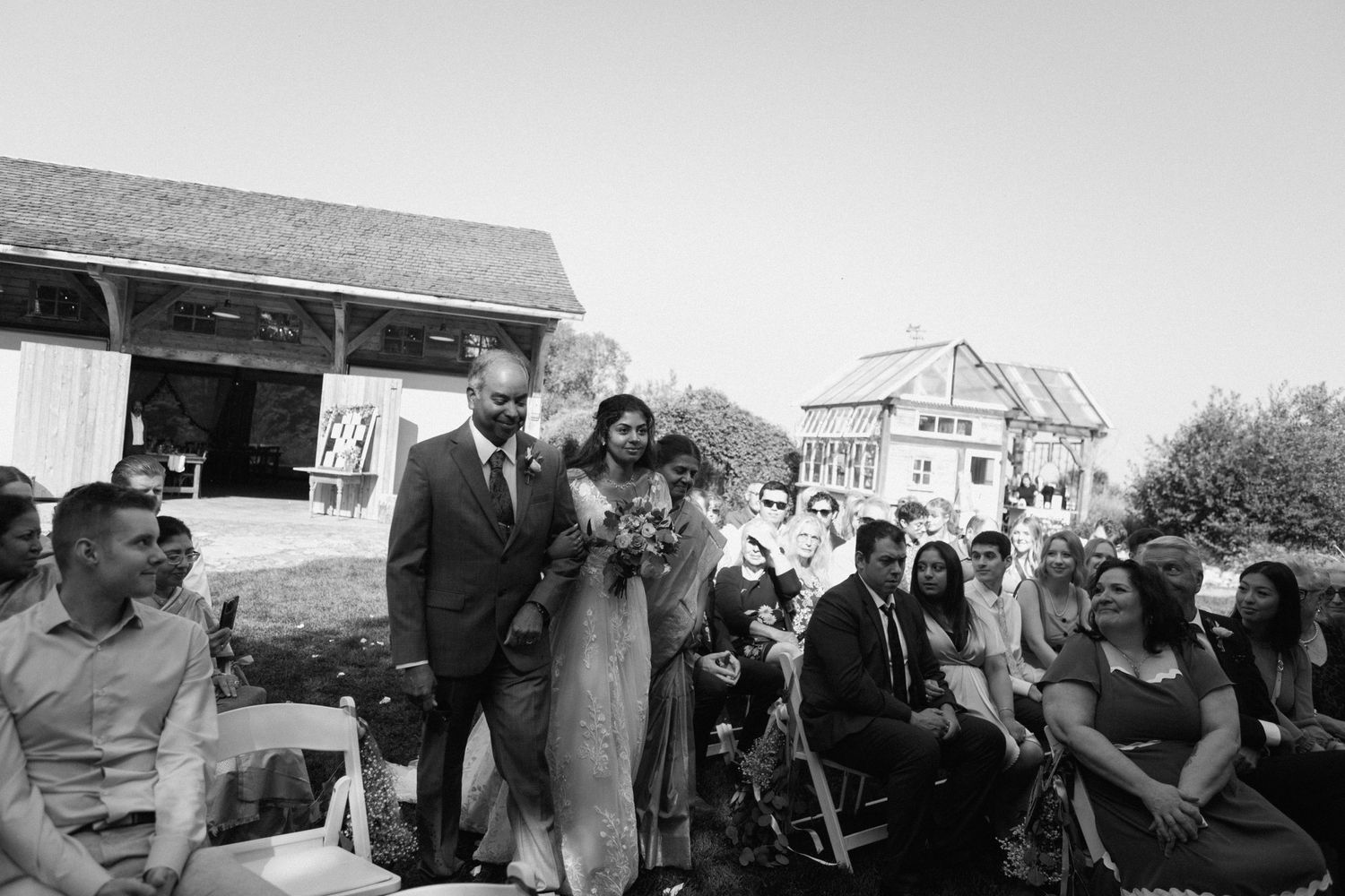 A black and white outdoor wedding ceremony with guests seated on benches at a rustic venue.