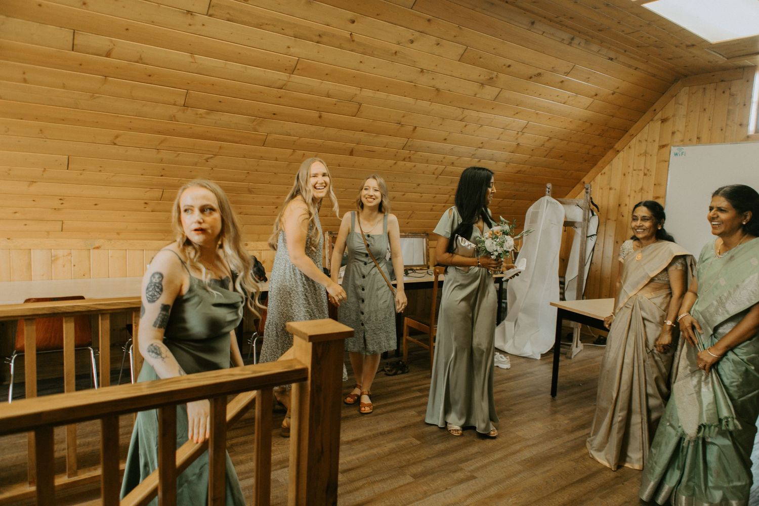 A group of people wearing coordinating gray and neutral bridesmaid dresses gather in a wooden-paneled room.
