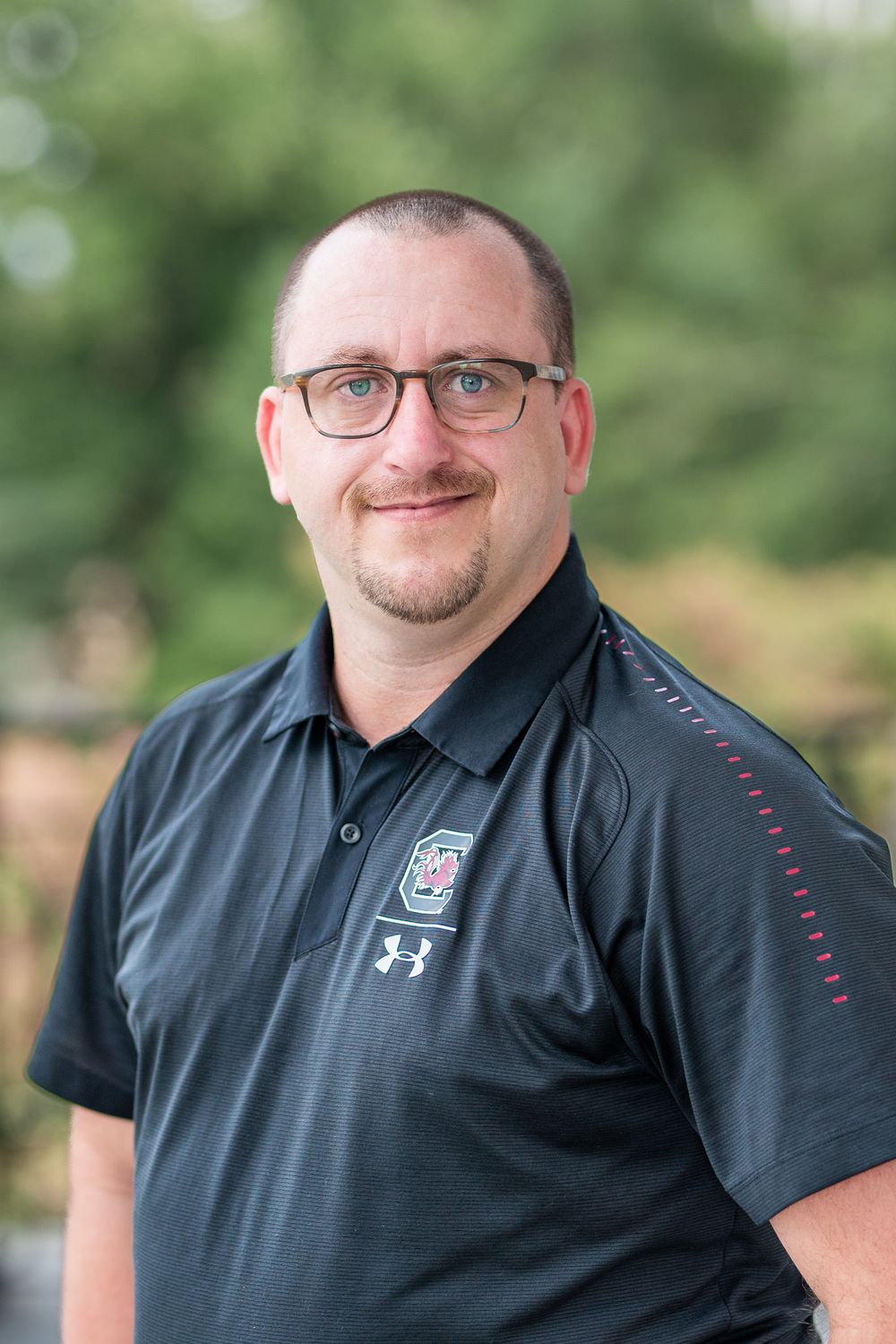 A person wearing glasses and a black Under Armour polo shirt smiles at the camera in an outdoor setting.