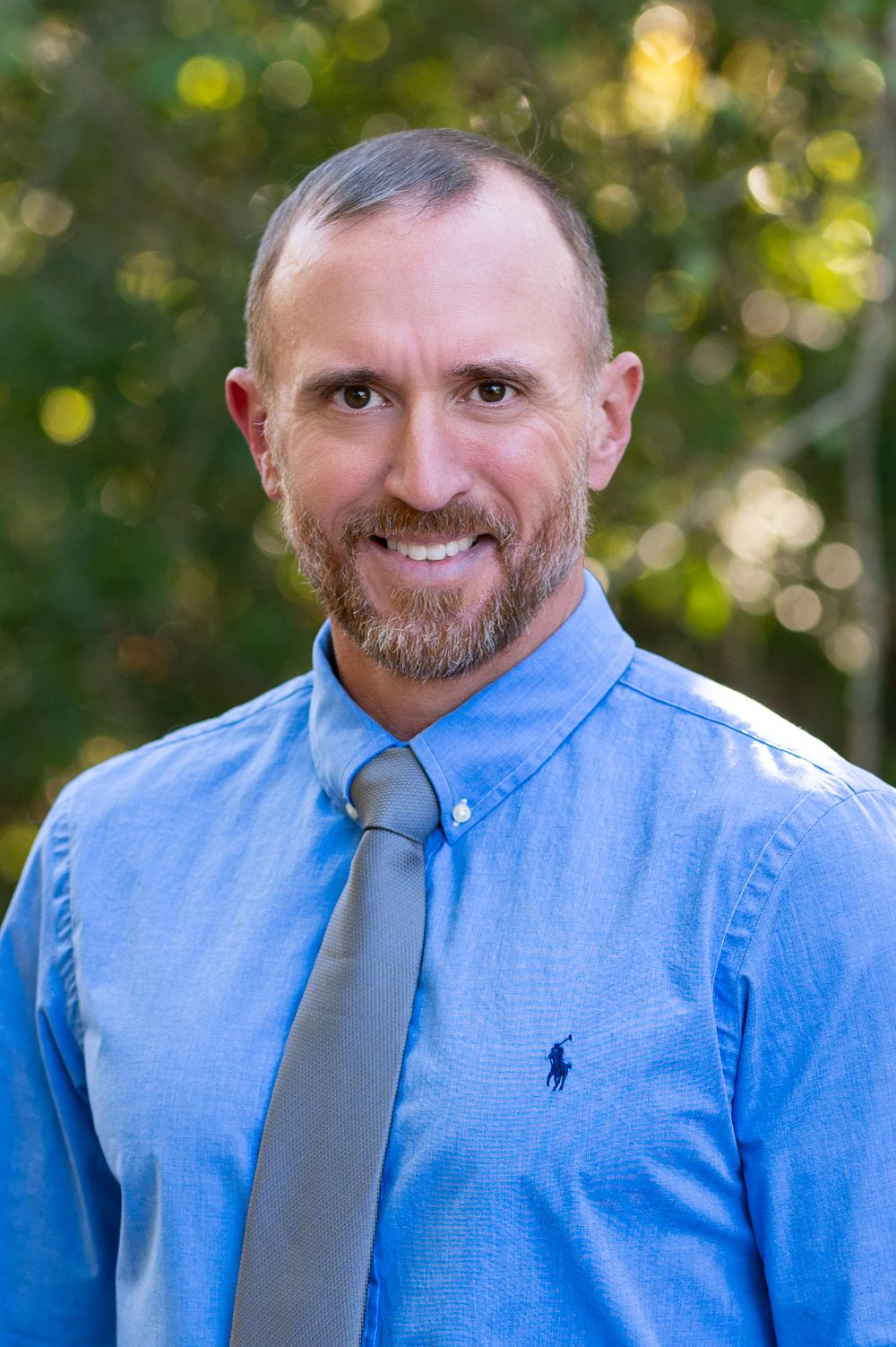 Professional in light blue button-down shirt and gray tie smiles against natural outdoor backdrop.