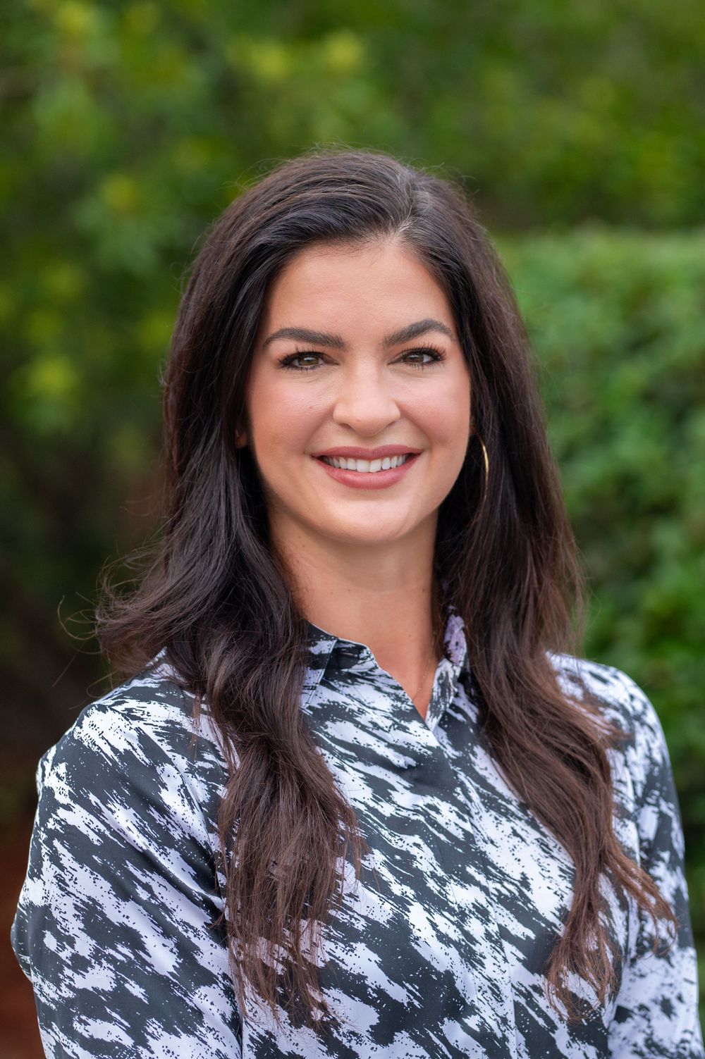 Professional headshot of person wearing a black and white patterned blouse against a green outdoor background.