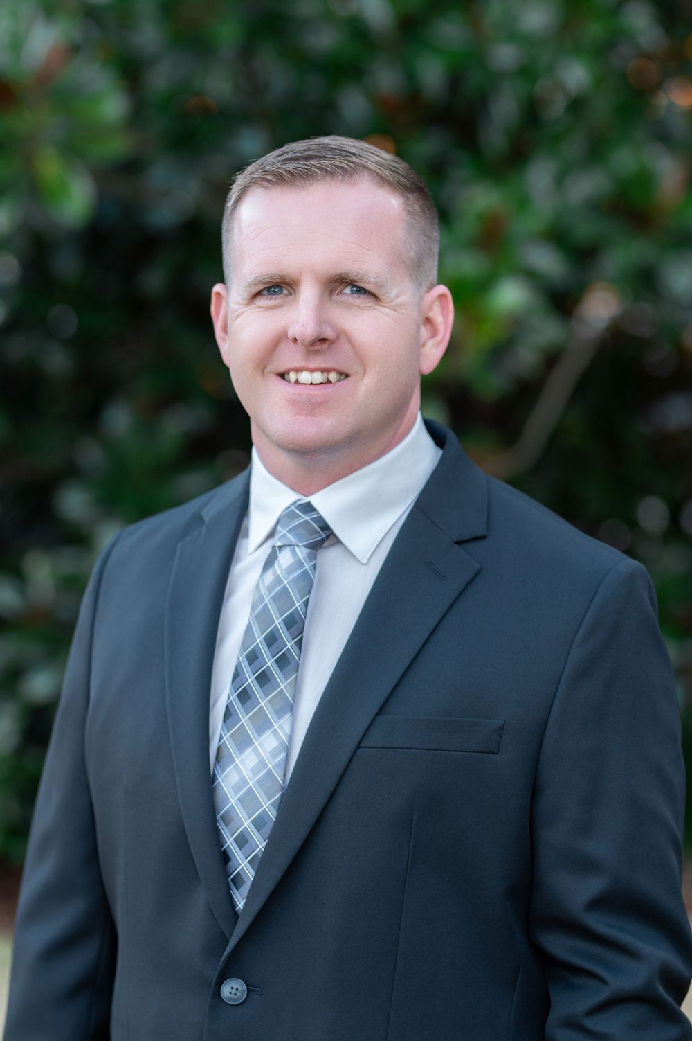 Professional headshot series of person in grey suit and plaid tie against greenery.