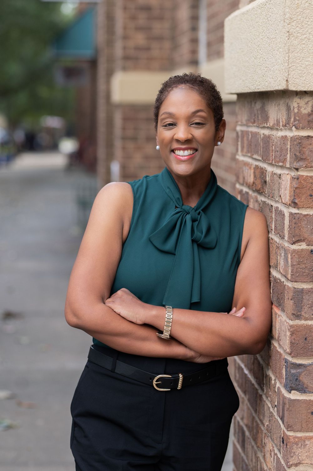 Business portrait of person in dark teal blouse leaning against brick wall outdoors.