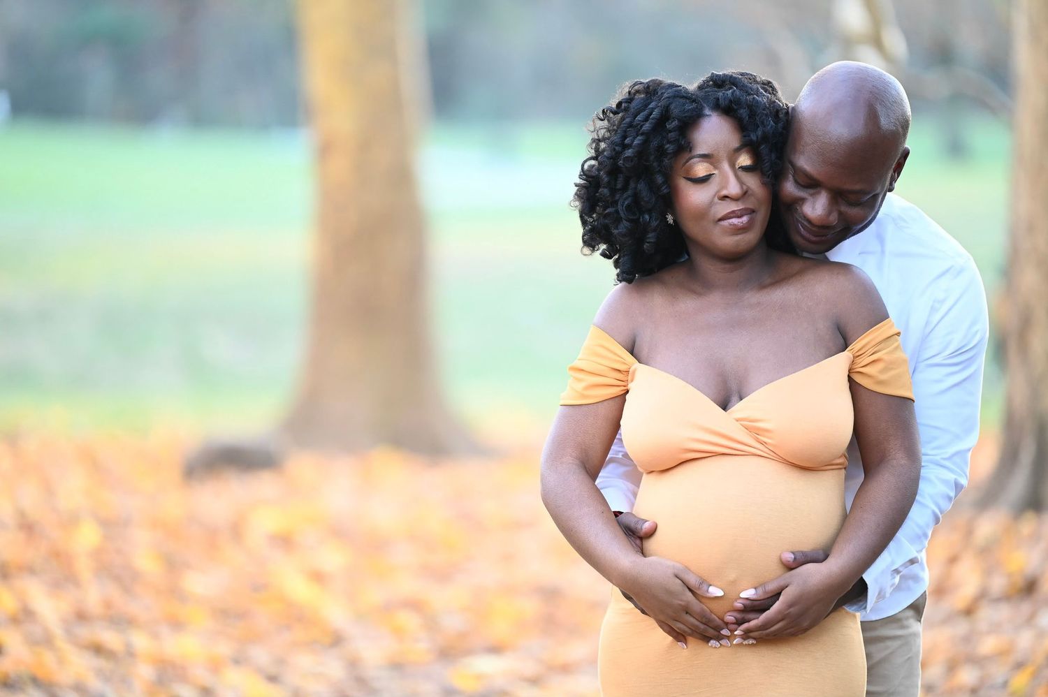 “A couple standing in a scenic outdoor setting, the man embracing his pregnant partner as they share a loving moment. Natural and emotional