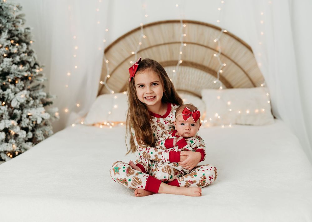 Child in festive pajamas sitting on bed with Christmas decorations and lights.