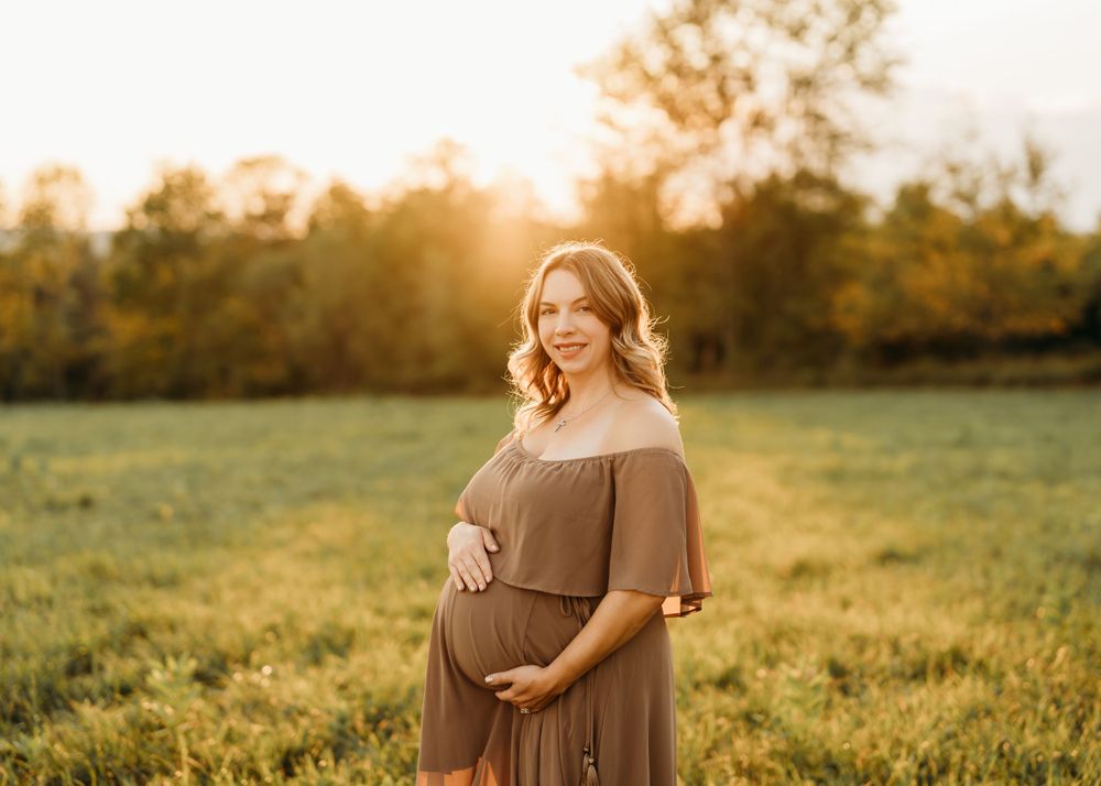 A maternity portrait in a brown dress against a sunlit meadow at golden hour.
