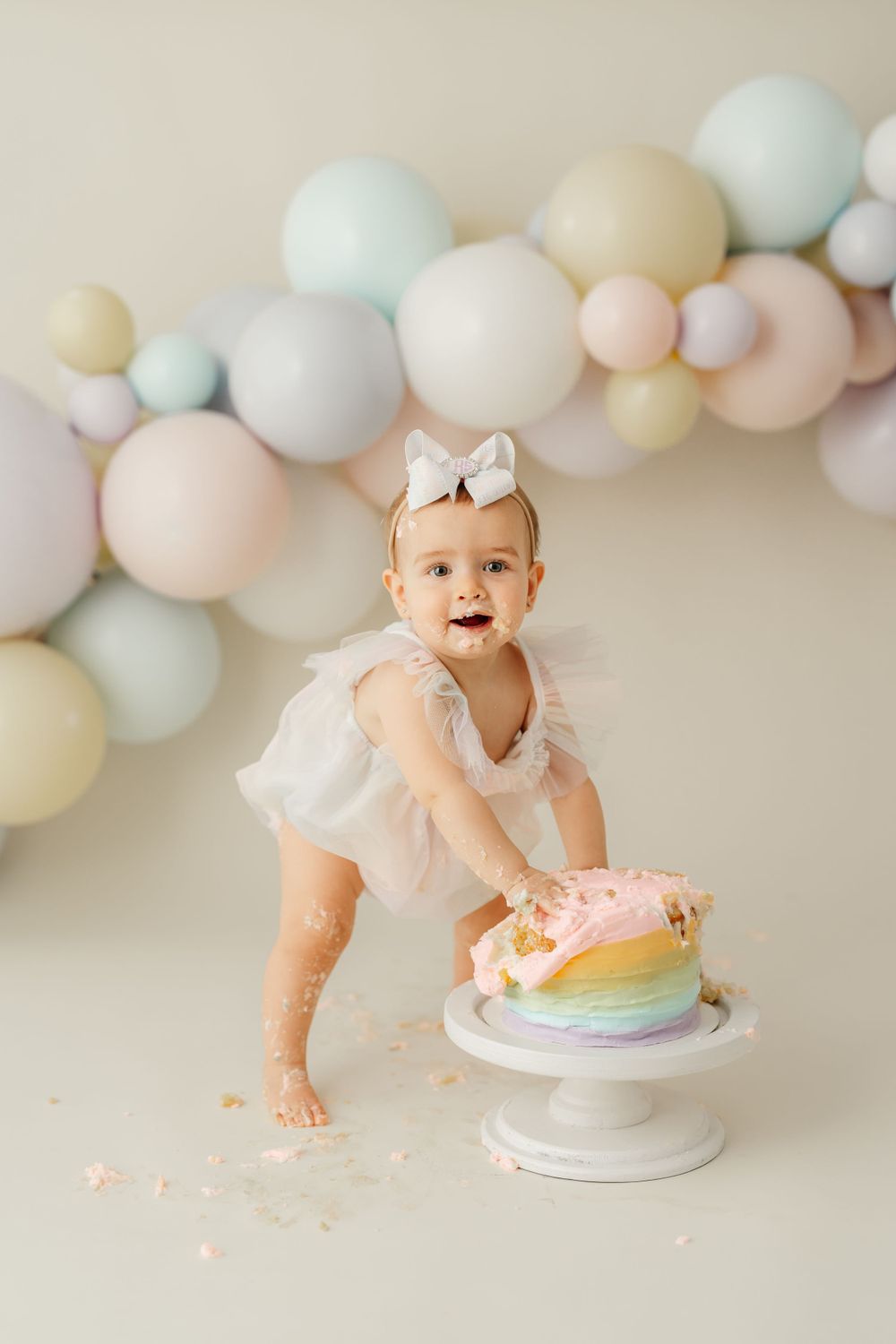 Baby in white dress reaching for rainbow cake with pastel balloon arch backdrop.